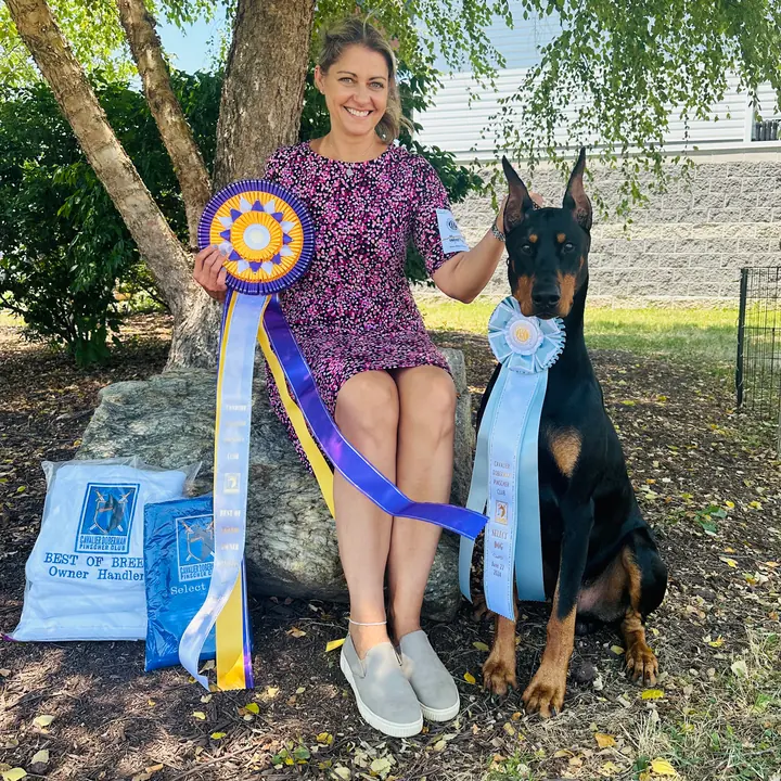 Kimberly Gabor Piorkowski seated outdoors with a Doberman Pinscher and ribbons